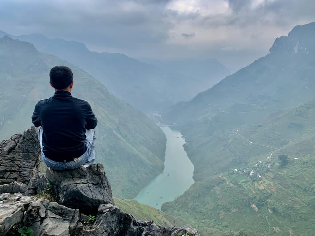 Asian male sitting on rocky peak overlooking Ma Pi Leng Pass in Ha Giang Loop