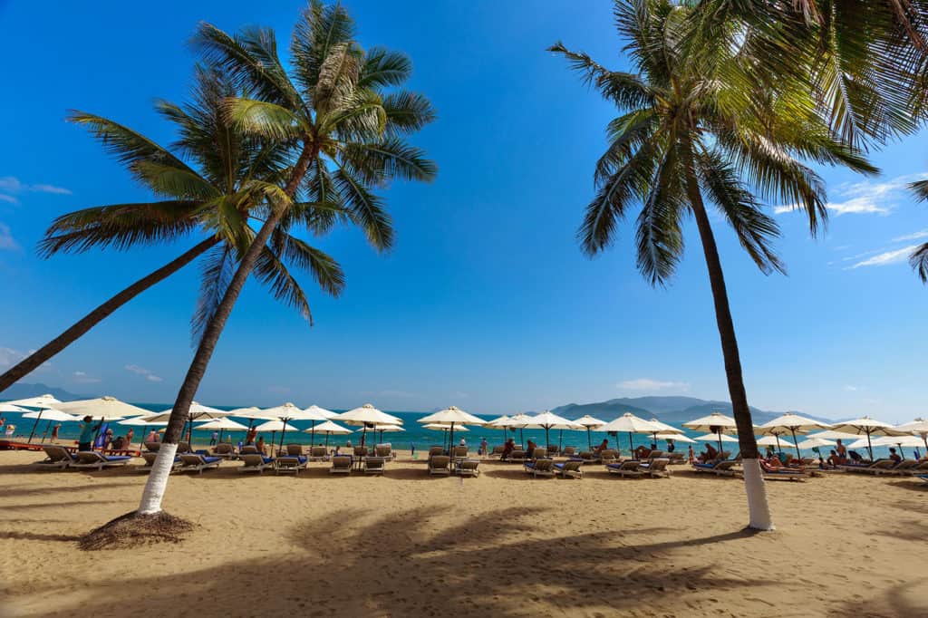 Beach in Nha Trang City, Vietnam as people relax under palm trees and lounge under beach umbrellas in the sun.